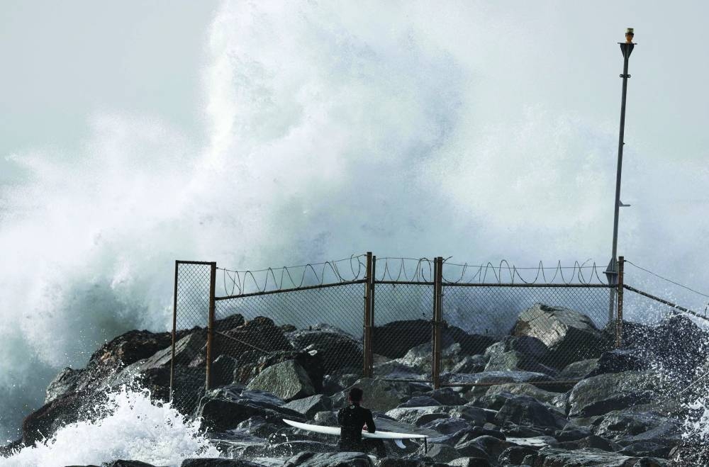 A surfer waits on a jetty on his way to enter the water as a large wave crashes near the beach in El Segundo, California.