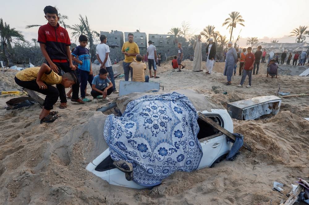 Palestinian boys stand near a buried damaged vehicle at the site following Israeli strikes on a tent camp sheltering displaced people at the Al-Mawasi area in Khan Younis, in the southern Gaza Strip, on Tuesday. REUTERS
