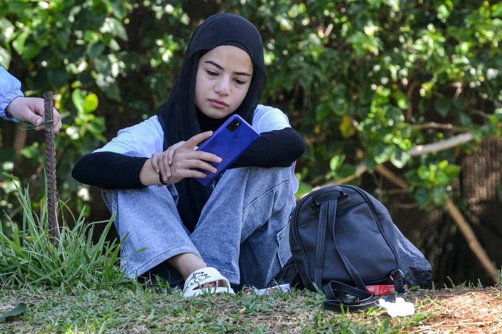 A girl browses a phone while sitting on a lawn at a public park as people displaced by conflict from southern Lebanon wait to be relocated to a temporary reception shelter in the northern city of Tripoli on Wednesday. AFP