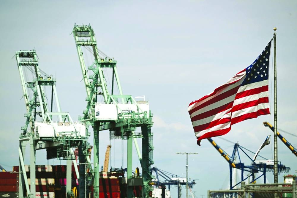 The US flag flutters in the wind as cranes stand above cargo shipping containers at the Port of Los Angeles, California.