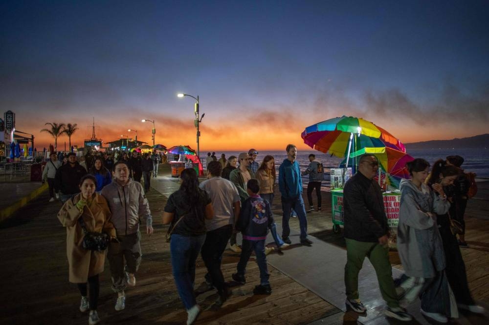 People walk at the Santa Monica pier during the sunset with smoke from the Palisades Fire seen in the sky on Saturday in Santa Monica, California. AFP