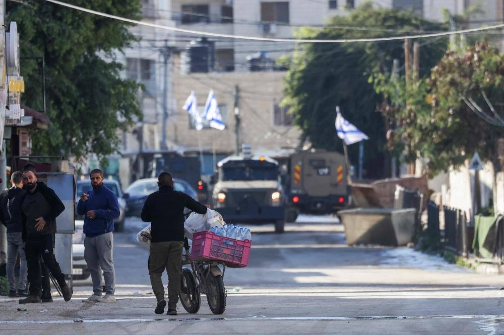 A Palestinian man pushes his bicycle loaded with supplies as Israeli forces in armoured vehicles conduct a raid in Jenin in the occupied West Bank, on Tuesday. REUTERS