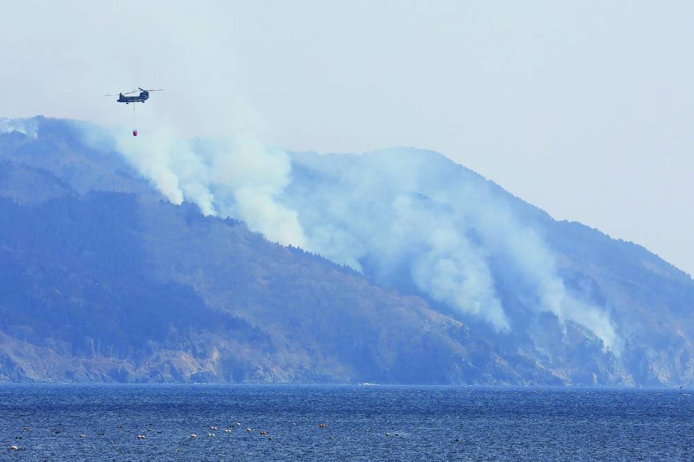 
A helicopter is pictured as smoke rises due to a wildfire on a mountainside near the city of Ofunato, Iwate Prefecture yesterday. At least one person has died in a wildfire that damaged more than 80 buildings and forced the evacuation of hundreds of residents in Japan, authorities said on Thursday. (AFP) 
