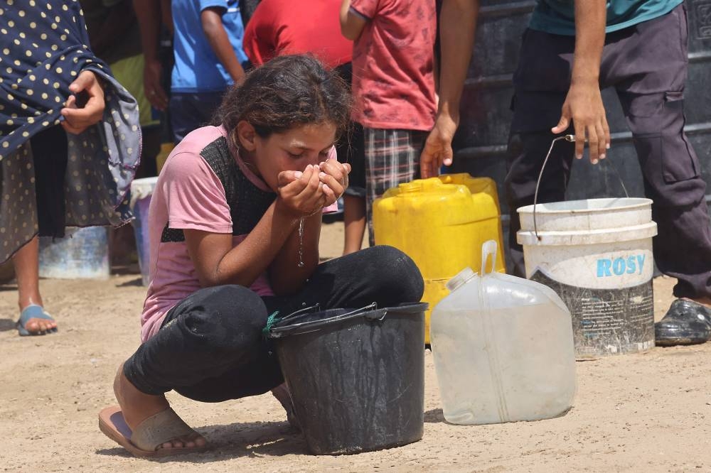 A girl drinks water from a plastic bucket while displaced Palestinians collect water in the Mawasi area of Khan Yunis, on August 14, 2025 (AFP)