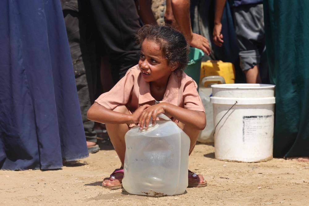 A girl squats by a water container while displaced Palestinians wait to collect  water in the Mawasi area of Khan Yunis, on August 14, 2025 (AFP)