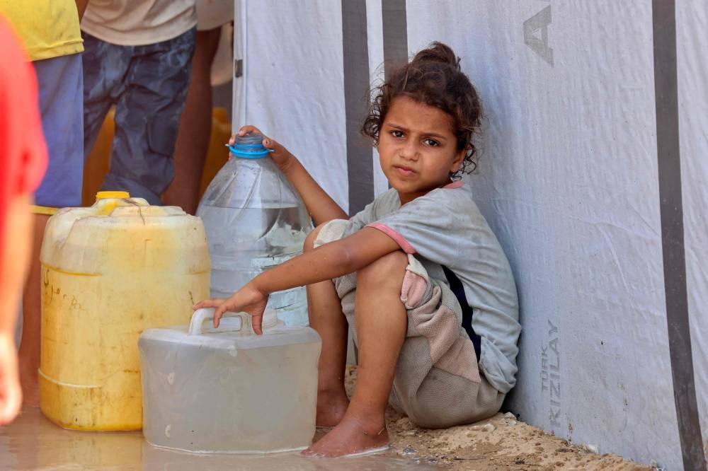 A girl sits by water containers while displaced Palestinians wait to collect water in the Mawasi area of Khan Yunis, on August 14, 2025 (AFP)