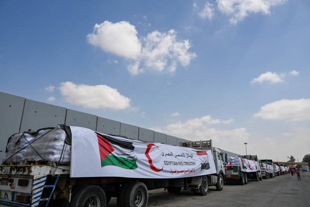 People walk past trucks loaded with aid for Gaza, waiting on the Egyptian side of the Rafah crossing in Rafah on August 18, 2025. Rights group Amnesty International on August 18 accused Israel of enacting a "deliberate policy" of starvation in Gaza, as the United Nations and aid groups warn of famine in the Palestinian territory. Israel, while heavily restricting aid allowed into the Gaza Strip, has repeatedly rejected claims of deliberate starvation in the 22-month-old war. (AFP)