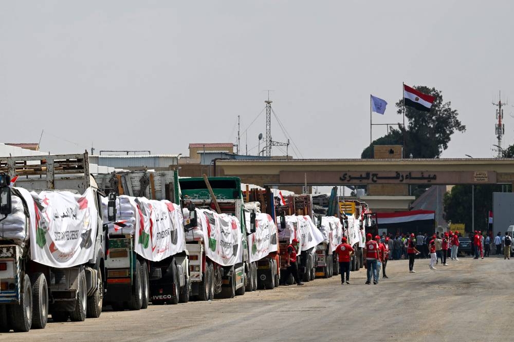 People walk past trucks loaded with aid for Gaza, waiting on the Egyptian side of the Rafah crossing in Rafah on August 18, 2025. Rights group Amnesty International on August 18 accused Israel of enacting a "deliberate policy" of starvation in Gaza, as the United Nations and aid groups warn of famine in the Palestinian territory. Israel, while heavily restricting aid allowed into the Gaza Strip, has repeatedly rejected claims of deliberate starvation in the 22-month-old war. (AFP)