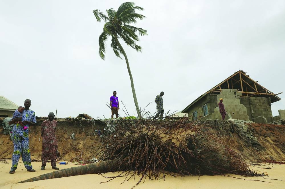 People stand near an uprooted coconut tree marking the spot where their home once stood before it was swept away by ocean surges, in Okegelu, Nigeria.