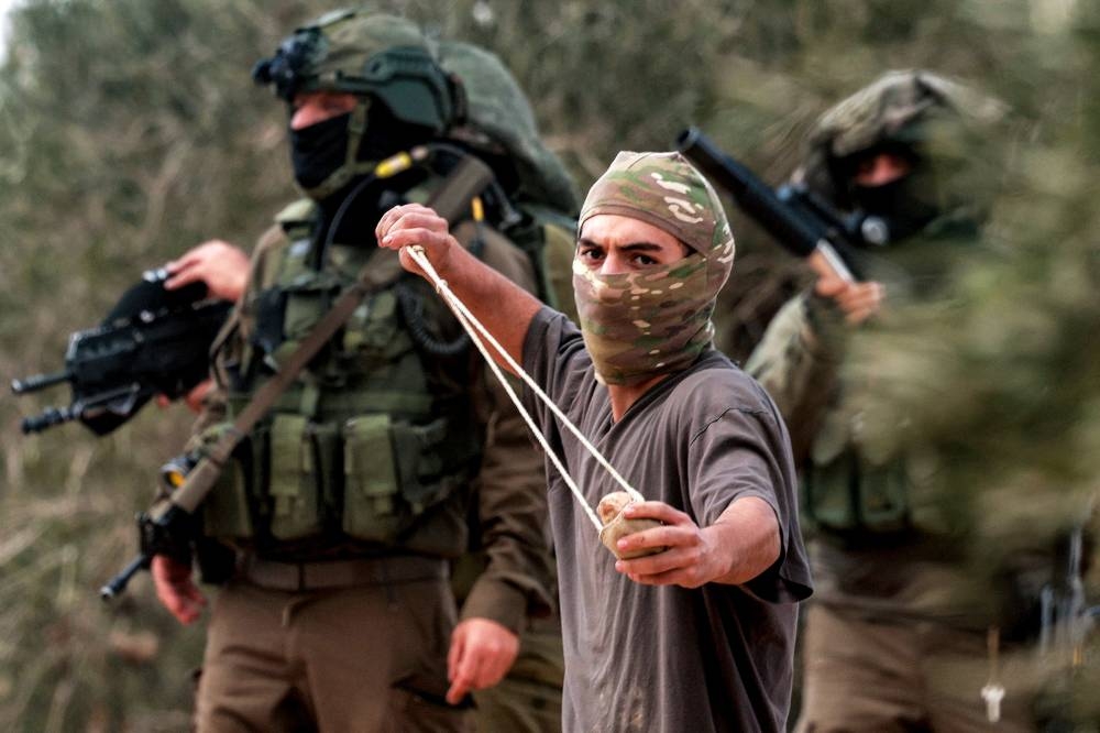 Israeli army soldiers stand behind a masked man swinging a slingshot while hurling stones at Palestinians who had gathered for the annual olive harvest season, during an attack by Israeli settlers on the Palestinian village of Beita, south of Nablus in the occupied West Bank, on October Friday. Agence France-Presse's (AFP) photographer Jaafar Ashtiyeh who took this photo was injured in the attack by Israeli settlers on October 10 while covering the olive harvest. 