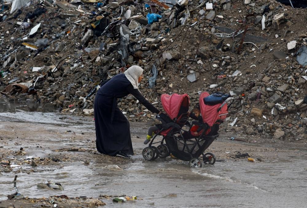 A Palestinian woman pulls a stroller with children, next to piles of rubble, during a rainy day in Gaza City, November 25, 2025. REUTERS