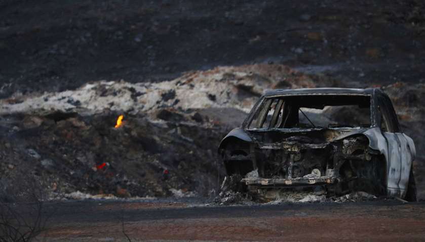 The burnt wreckage of a vehicle is seen along a road as a flames are seen, in the aftermath of the W