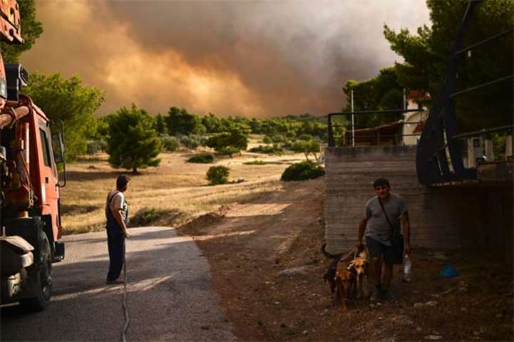 A man walks his dog past another man holding a hose as smoke billows in background in Kineta