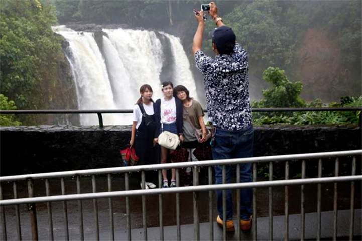 A guide takes pictures of tourists in front of a waterfall swollen by rain from Hurricane Lane