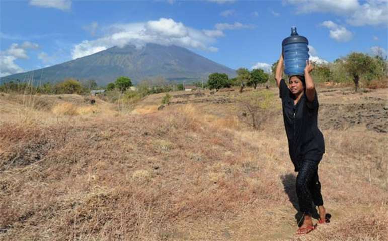 A woman carries water on her head, as Mount Agung volcano looms in the background