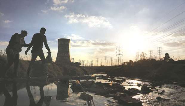 A couple crosses a stream flowing towards electricity pylons and the cooling tower of the defunct Orlando Power Station in Soweto, South Africa (file). More than a decade of unreliable supply and surging prices are driving consumers and businesses off the grid as the price of renewable energy drops, leaving Eskom with lower sales and high fixed costs due to the expense of building new power plants.