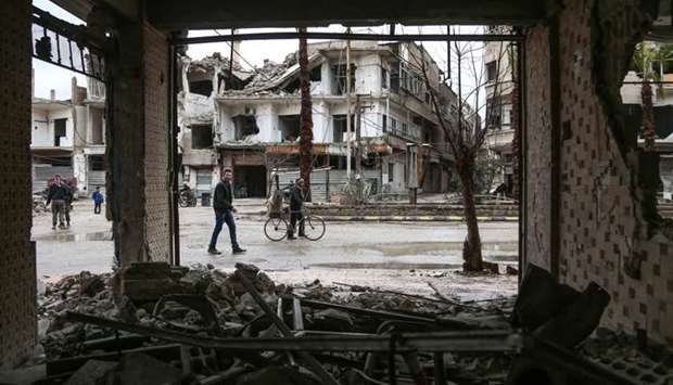 A picture taken on February 17, 2018 shows a man walking next to another pushing a bicycle loaded with a carpet, down a rain-soaked street past damaged and destroyed buildings in the Syrian rebel-held enclave of Arbin in the Eastern Ghouta near the capital Damascus.