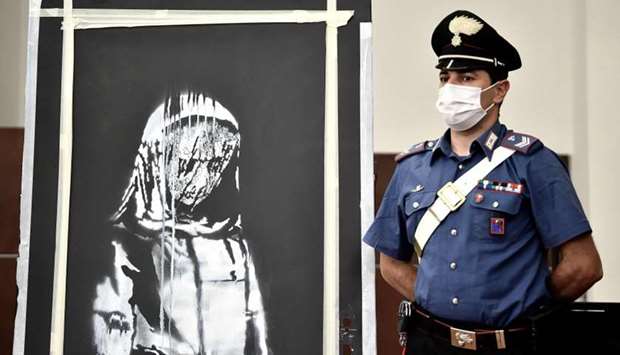 In this file photo taken on June 11, 2020, an Italian Carabinieri poses near a piece of art attributed to Banksy, that was stolen at the Bataclan in Paris in 2019, and found in Italy, during a press conference in L'Aquila