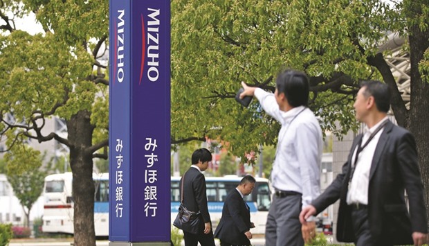 Pedestrians walk past signage for Mizuho Bank displayed outside a branch in Tokyo. Mizuho Financial Group, the fourth-largest lender to Saudi Arabia, is expanding advisory services in the kingdom, as banks jockey for a role in Saudi Aramcou2019s expected $100bn initial public offering.